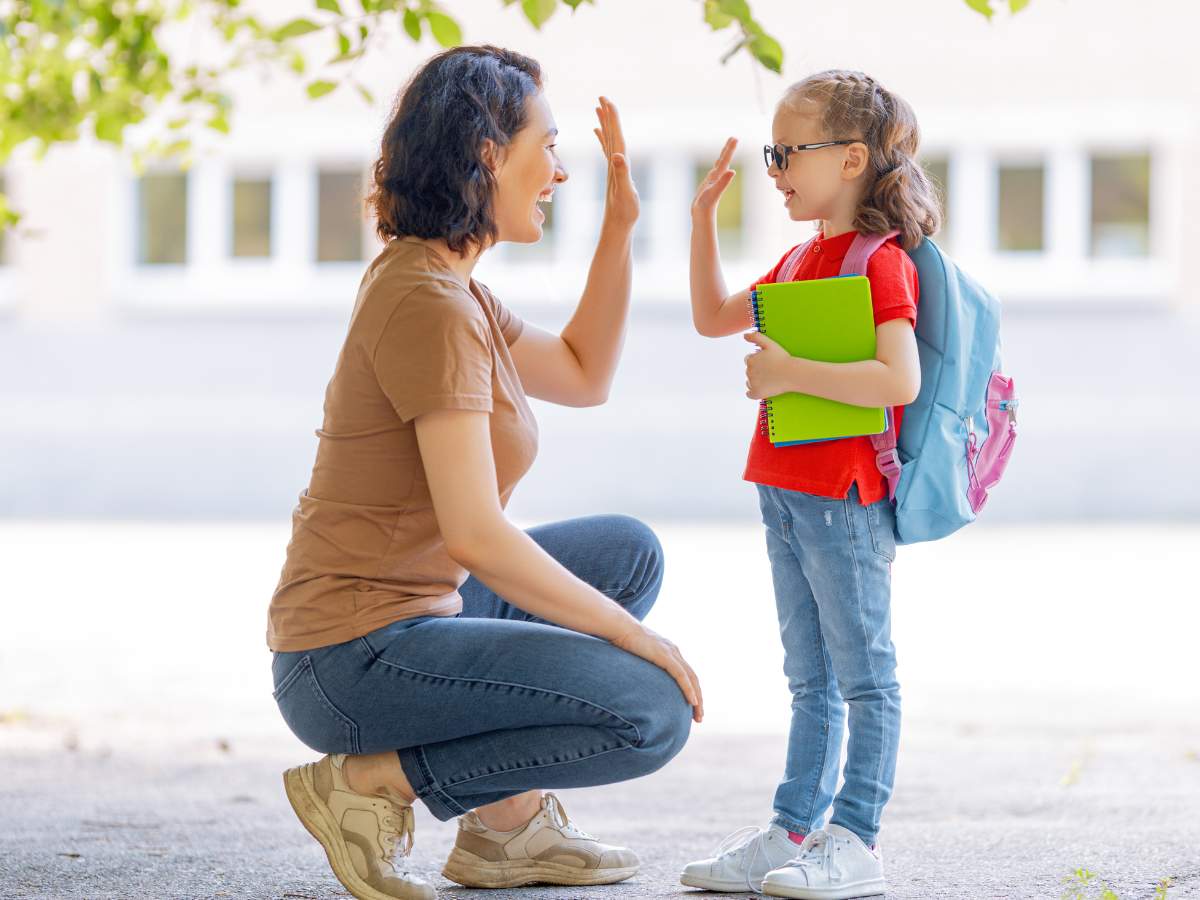 An adult and a child wearing a backpack and holding a notebook exchange a high-five outdoors.