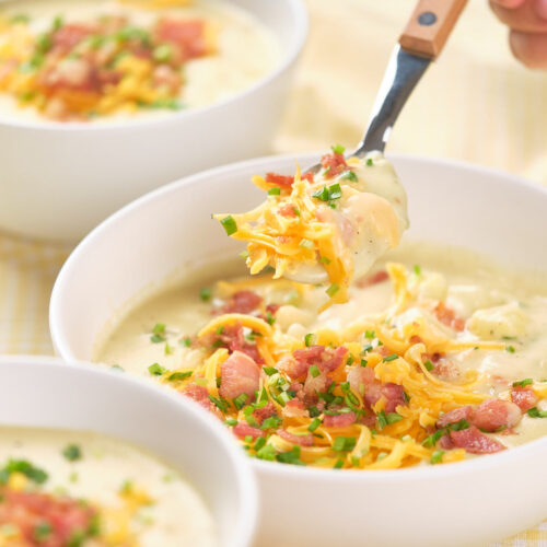 A spoonful of baked potato soup being lifted from a white bowl filled with the same soup.