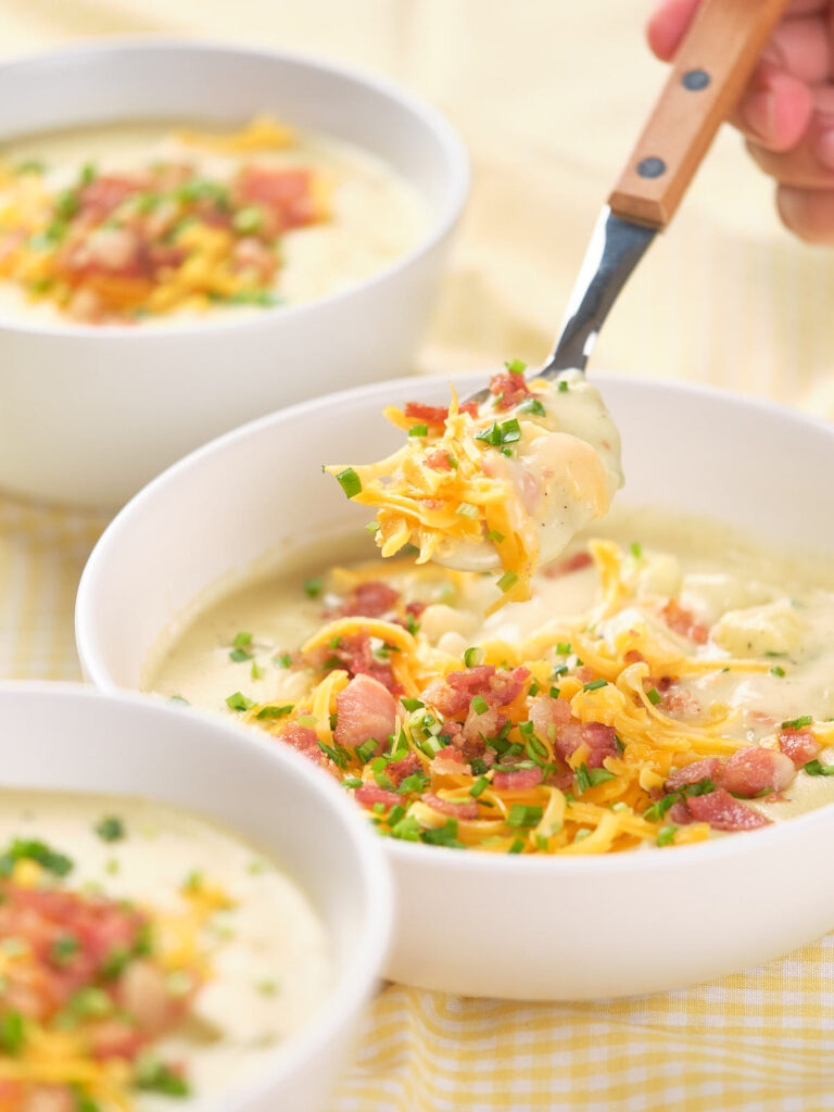 A spoonful of baked potato soup being lifted from a white bowl filled with the same soup.