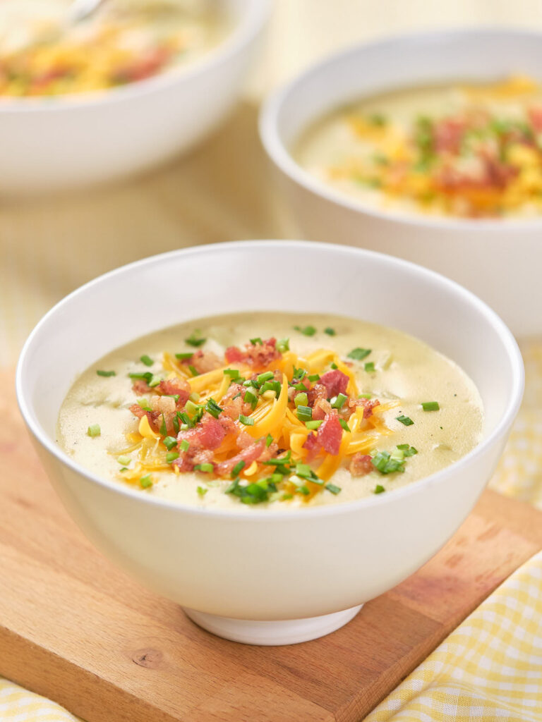 A bowl of baked potato soup garnished placed on a wooden surface with two similar bowls in the background.