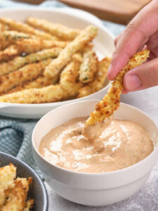 A hand dipping a fried zucchini fry into a blackened ranch sauce.