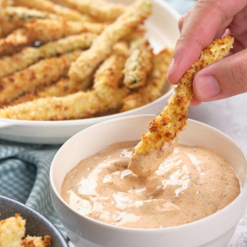 A hand dipping a fried zucchini fry into a blackened ranch sauce.