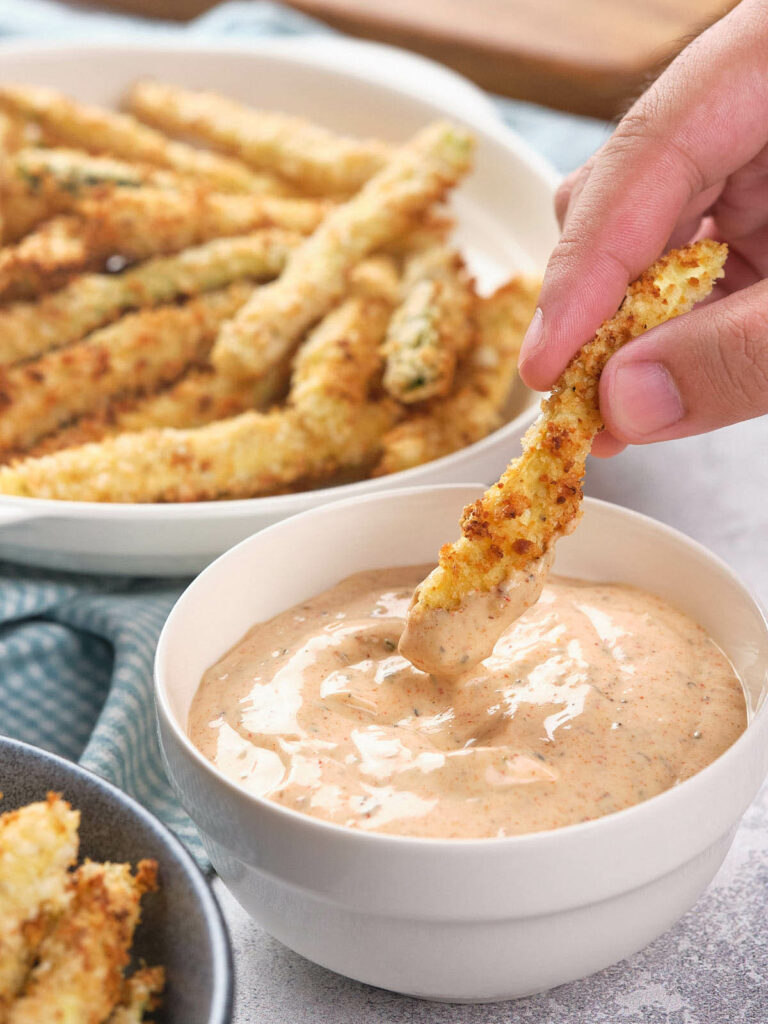 A hand dipping a fried zucchini stick into a creamy dipping sauce.