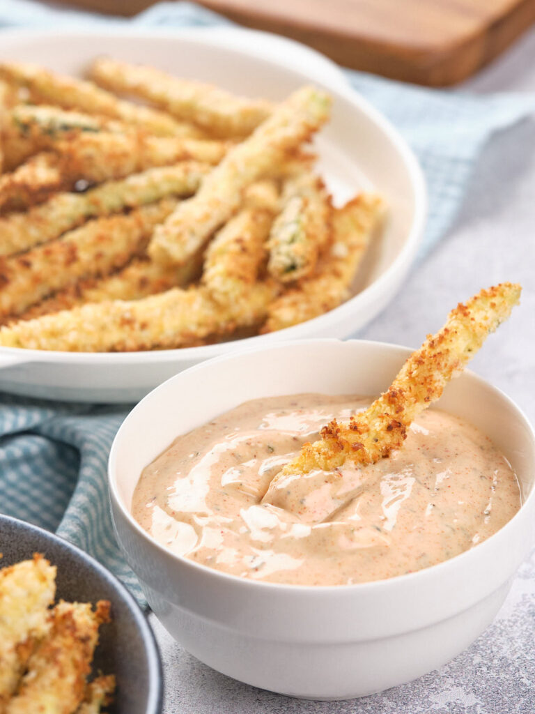 A bowl of creamy dip with a fried zucchini stick dipped in it.