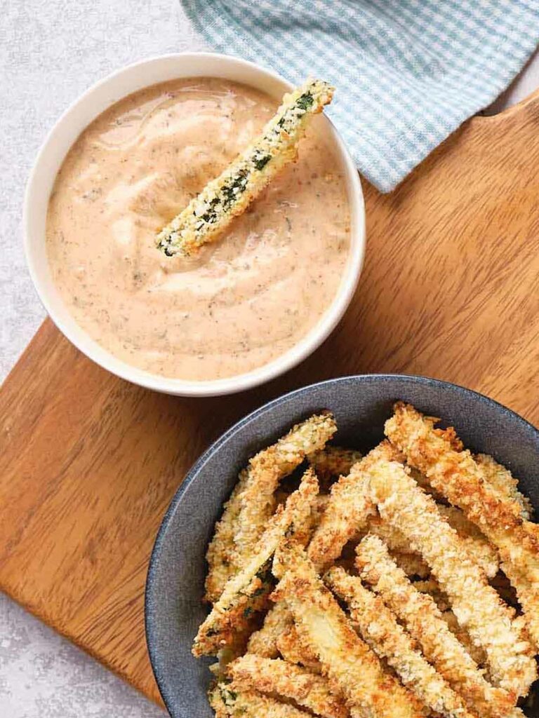 A bowl of crispy zucchini fries is placed next to a bowl of dipping sauce on a wooden surface.