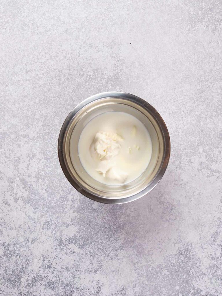 A small metal bowl containing thick white cream sits on a textured gray surface.