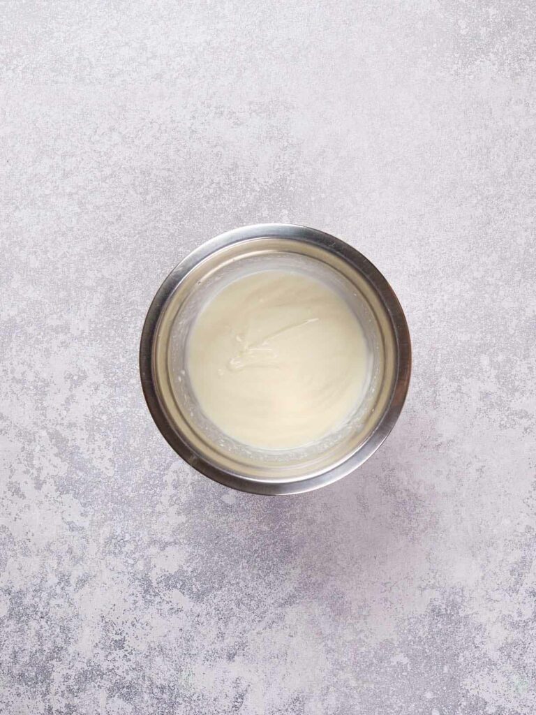 A small stainless steel bowl containing smooth, white cake dough is placed on a textured, light gray surface.