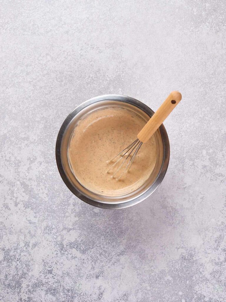 A metallic bowl filled with a thick, beige-colored mixture sits on a textured surface; a whisk with a wooden handle is inside the bowl.