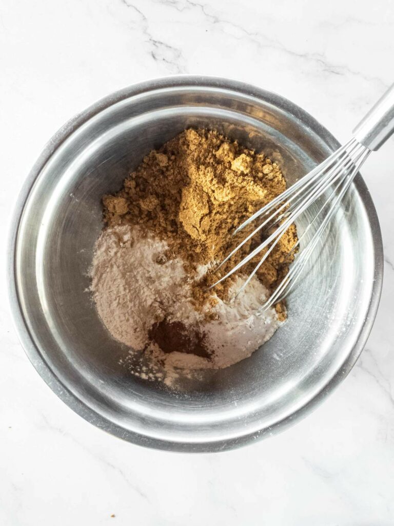 A stainless steel bowl containing brown sugar and other dry baking ingredients being mixed with a whisk.