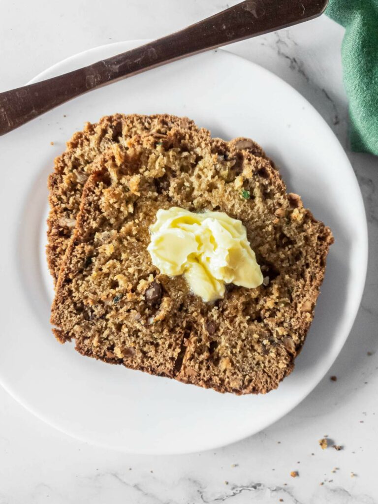 Two slices of zucchini bread with a dollop of butter are served on a white plate.