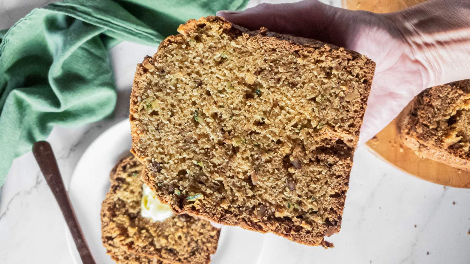 A hand holding a slice of zucchini bread, with a green cloth and knife visible in the background on a marble surface.