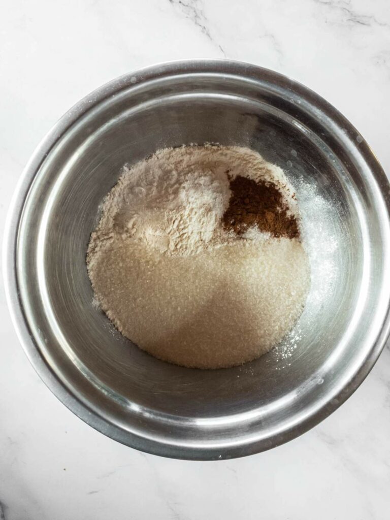 A metal bowl containing flour, sugar, and cinnamon powder on a marble countertop.