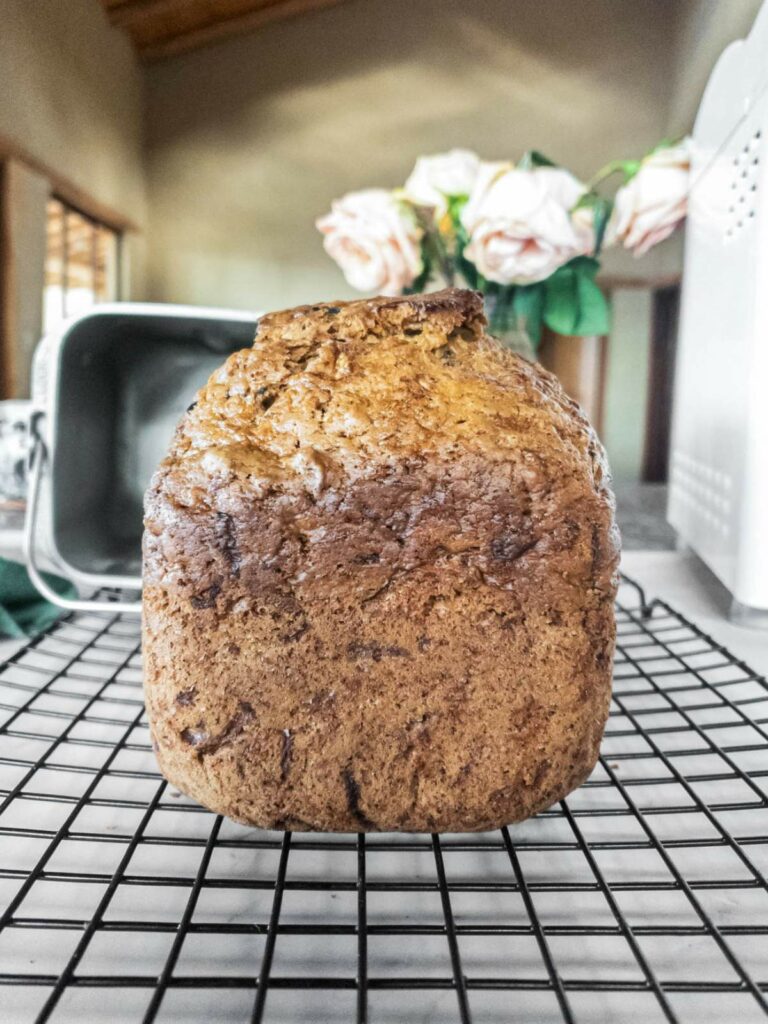 A freshly baked loaf of zucchini bread cooling on a wire rack with a bread machine and flowers in the background.