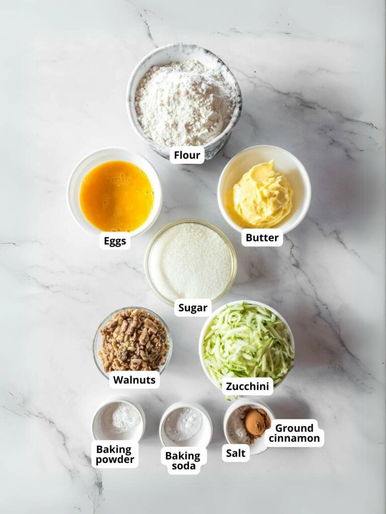 Ingredients of zucchini bread in bowls on a marble surface.