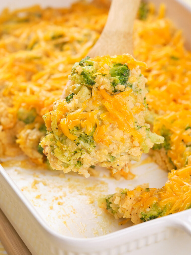 A wooden spoon lifting a serving of broccoli rice casserole topped with melted cheese from a white baking dish.