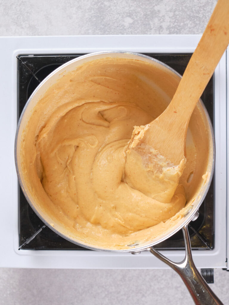 A pot of light brown batter is being stirred with a wooden spoon on a stovetop.