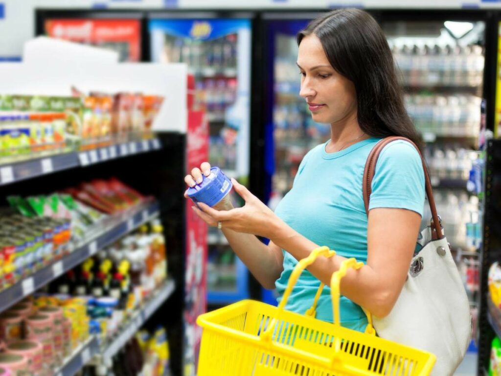 A woman stands in a grocery store aisle, holding a can and reading its label.