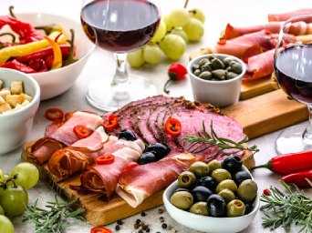 A charcuterie board with cured meats, olives, and red grapes, accompanied by two glasses of red wine, peppers, and bread cubes on a light-colored surface.