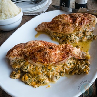 A white plate with two baked chicken breasts stuffed with a creamy spinach and cheese mixture. Mashed potatoes in a bowl and plates with cutlery are in the background.