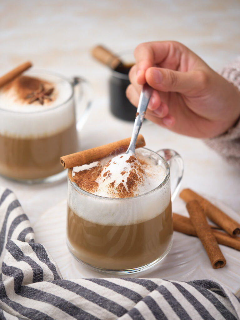 A hand stirs foam in a glass cup of dirty chai tea latte topped with ground cinnamon.