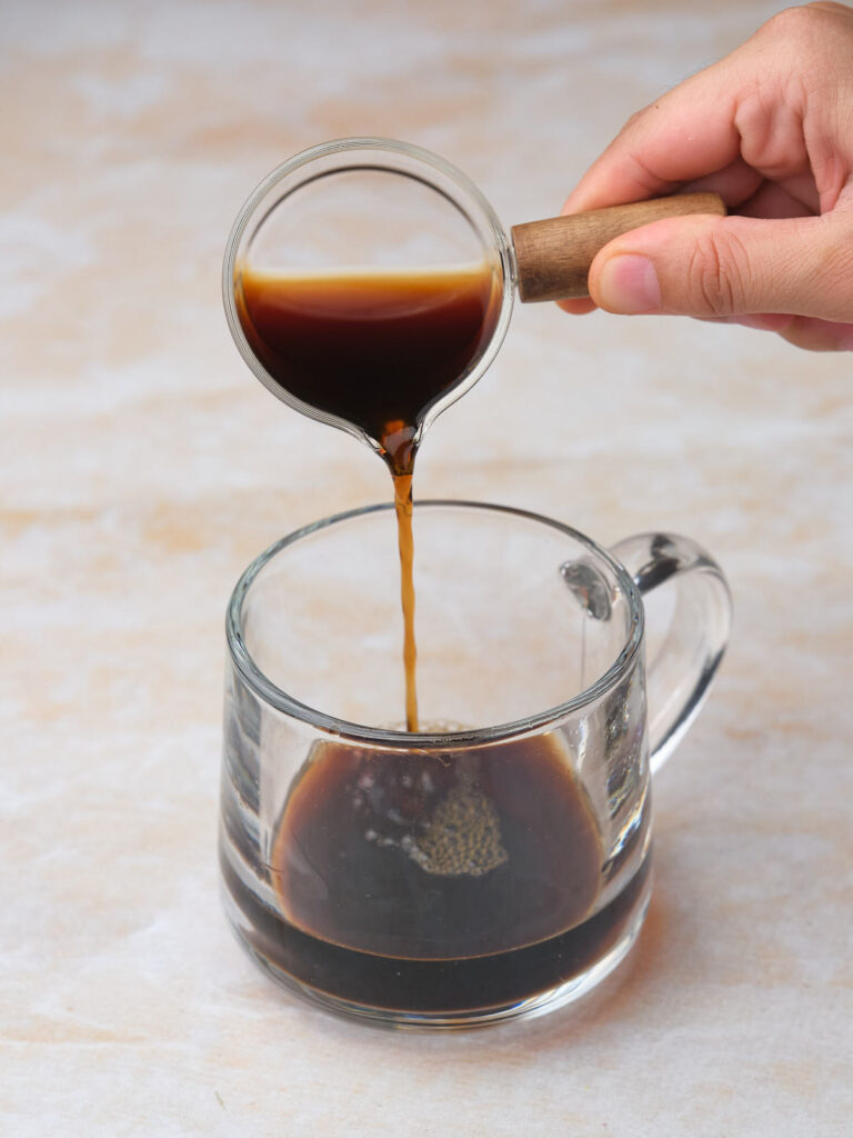 A hand is pouring coffee from a small glass container into a clear glass mug on a light-colored surface.