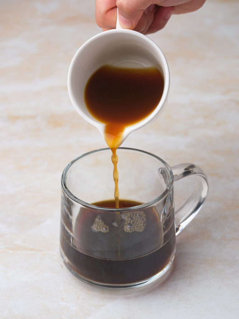 A hand pouring coffee from a white cup into a clear glass mug on a beige surface.