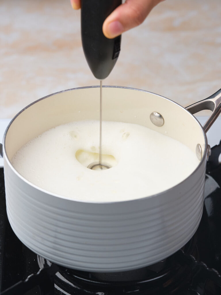 A hand uses a handheld frother to mix foamy milk in a pot on a stove.