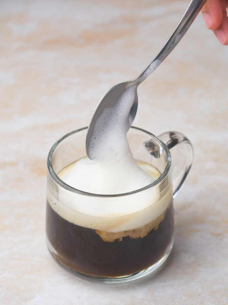 A spoon holding foam is being added to a clear glass mug of dirty chai tea latte.