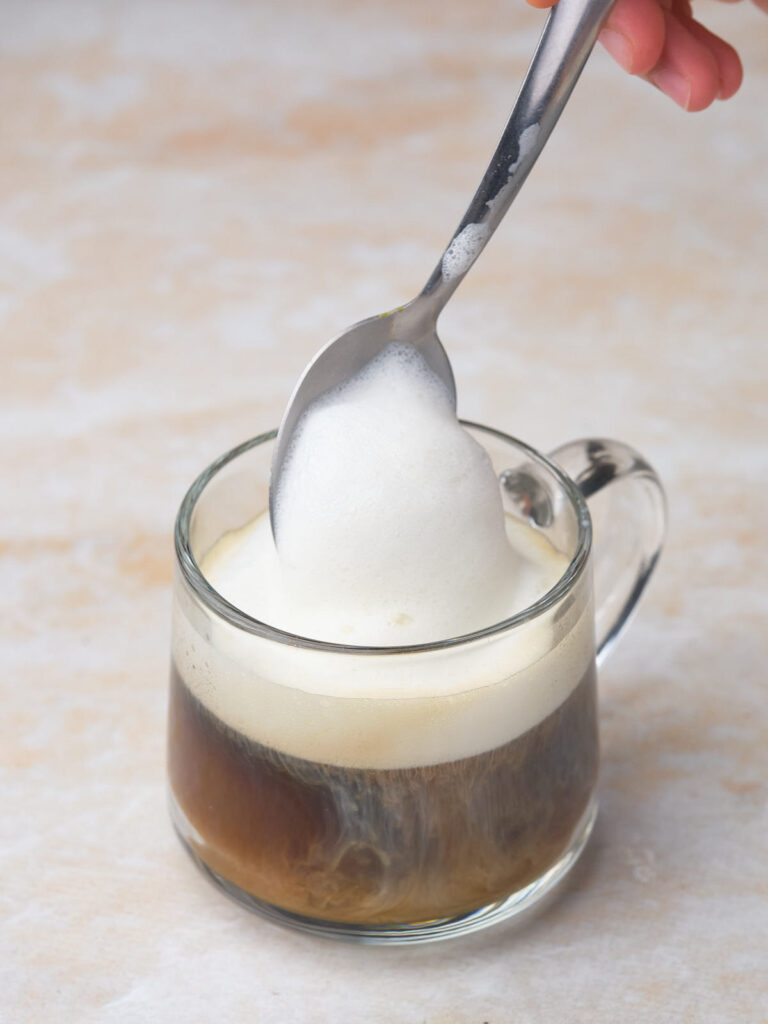 A hand is holding a spoon with frothy milk over a glass mug filled with dirty chai tea latte.