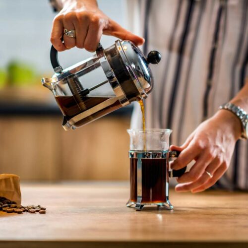 Person pouring coffee from a French press into a clear glass mug on a wooden table, with a bag of coffee beans nearby.