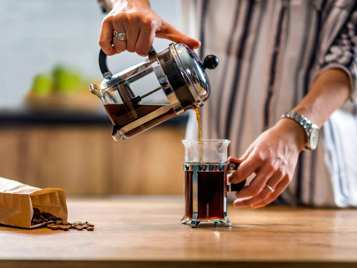 Person pouring coffee from a French press into a clear glass mug on a wooden table, with a bag of coffee beans nearby.