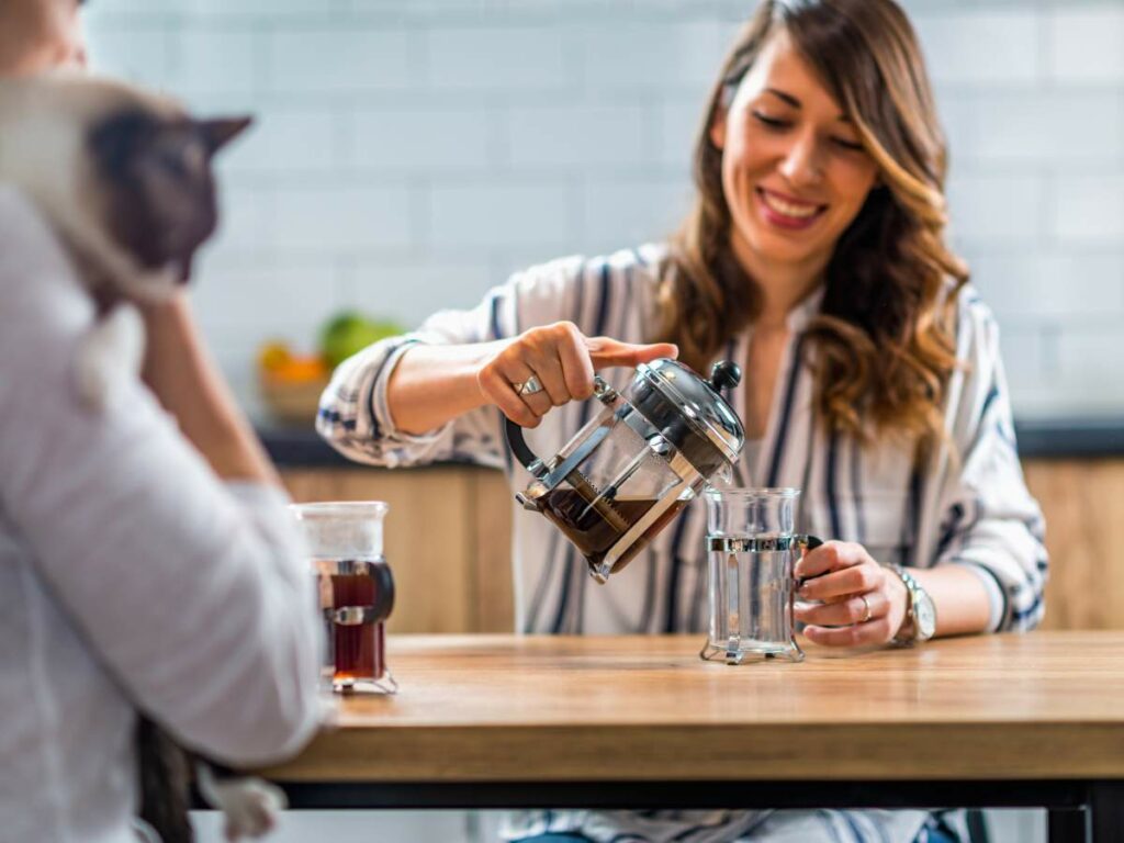 Two people at a kitchen table. One pours coffee from a French press into a glass.