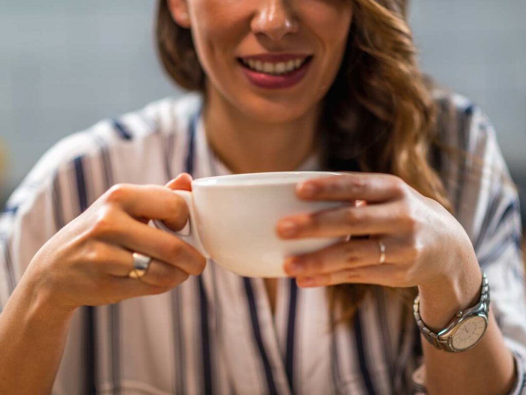 A person wearing a striped shirt holds a white cup with both hands while smiling.