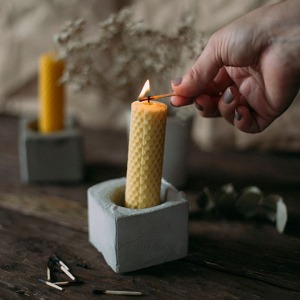 A hand is lighting a yellow candle with a match. The candle is in a white holder, and there are other candles and dried flowers in the background.