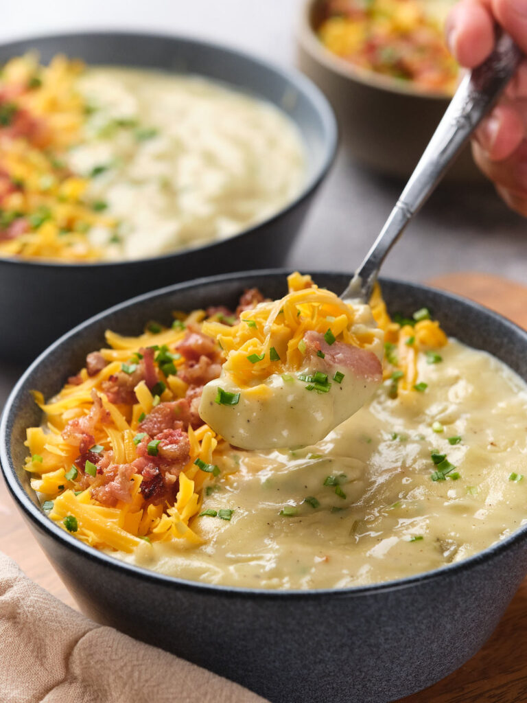 A person holds a spoonful of creamy potato soup with a bowl of the same soup in the background.