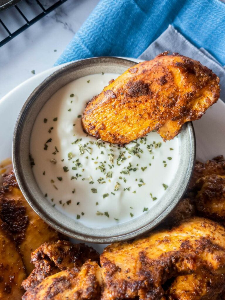 A piece of popeyes blackened chicken tenders being dipped into a bowl of creamy white sauce, garnished with green herbs.