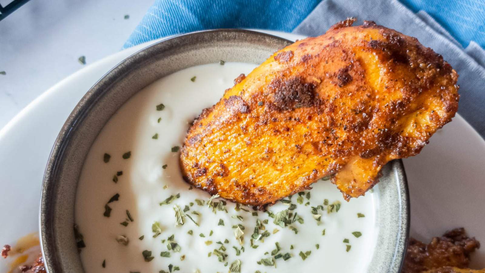 A crispy, seasoned chicken breast partially dipped into a bowl of creamy sauce.