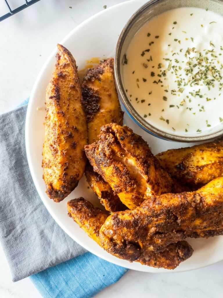 A plate of popeyes blackened chicken tenders next to a bowl of white dipping sauce garnished with herbs.