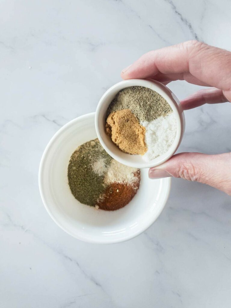 A hand holds a small bowl containing a mix of spices over a larger bowl.