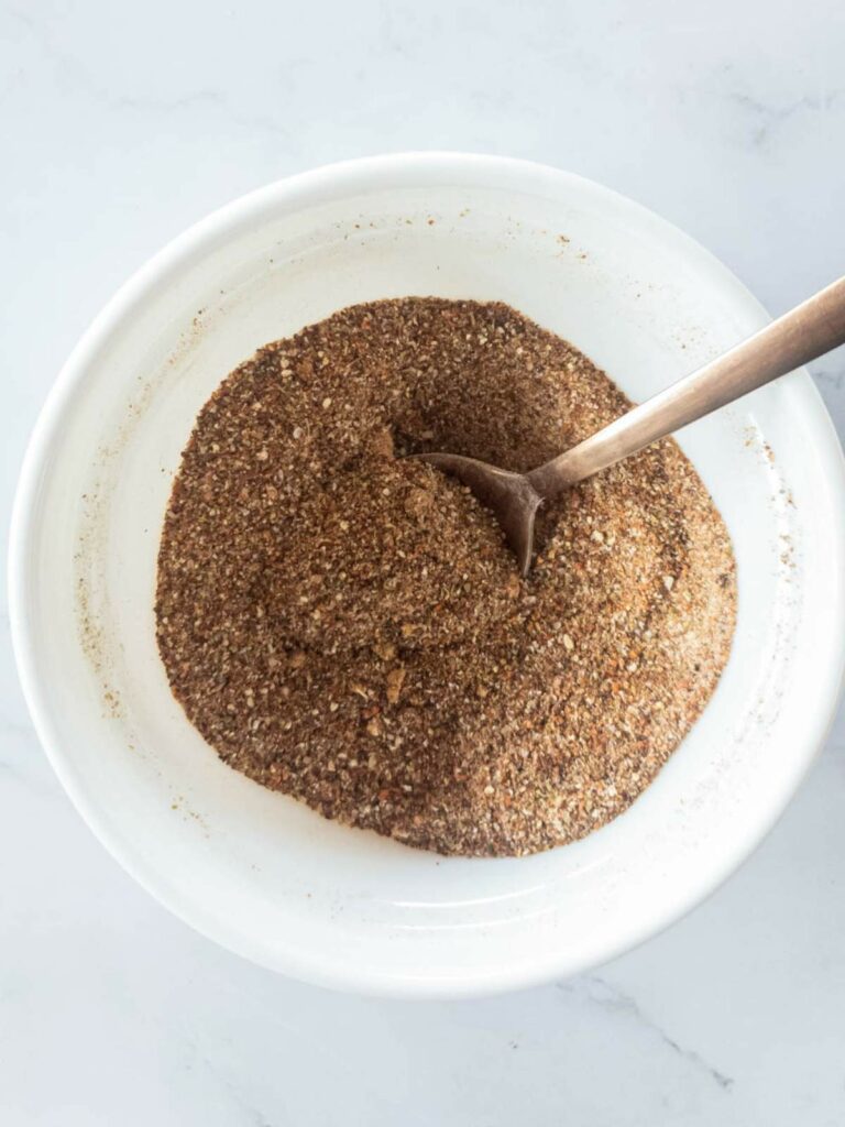 A white bowl filled with a finely ground brown seasoning mixture, with a spoon resting inside the bowl.