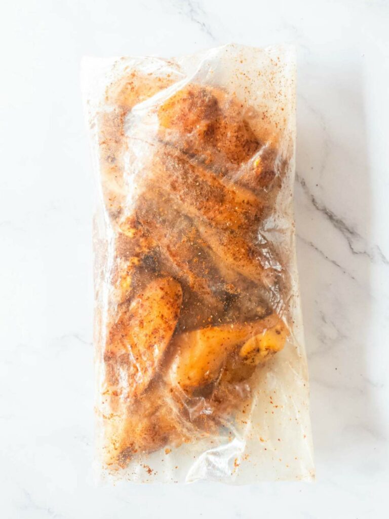 Chicken wings coated in seasoning, sealed in a plastic bag, placed on a white marble surface.