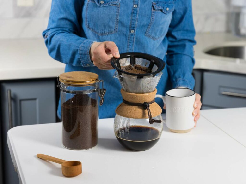 Person in a denim shirt brewing coffee using a pour-over coffee maker on a white countertop.