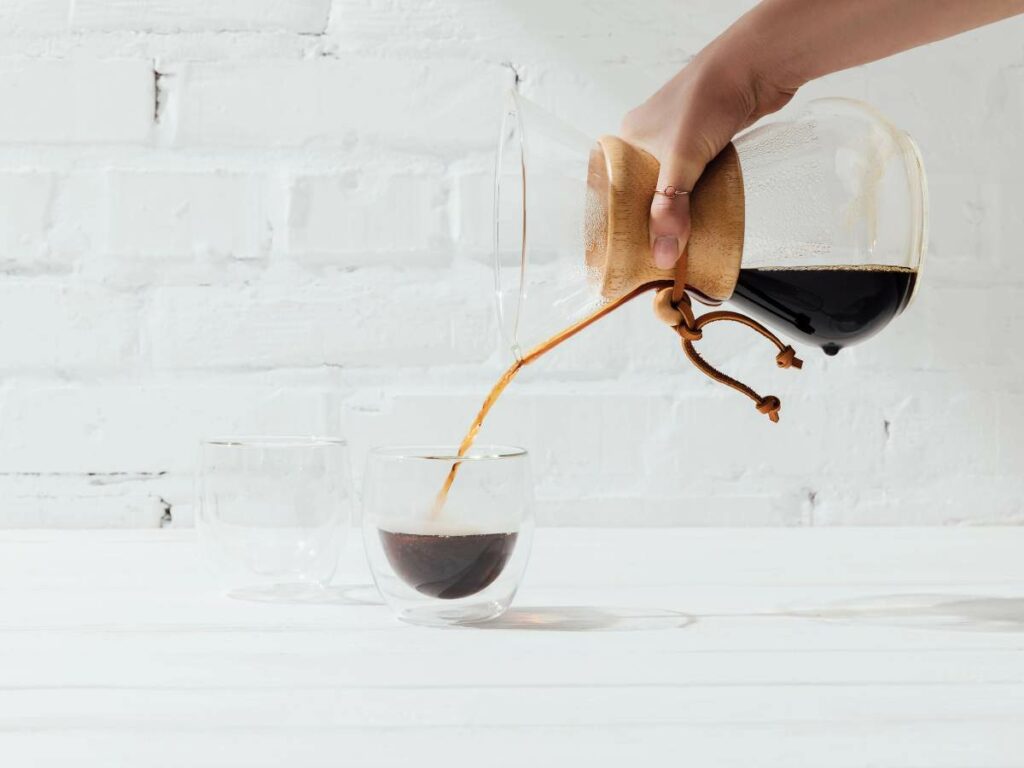 A hand pours coffee from a carafe into a glass cup on a white table with a brick wall background.