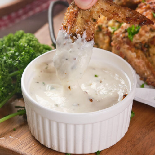 A hand dipping a seasoned chicken wing into a wingstop ranch dressing in a small white ramekin.