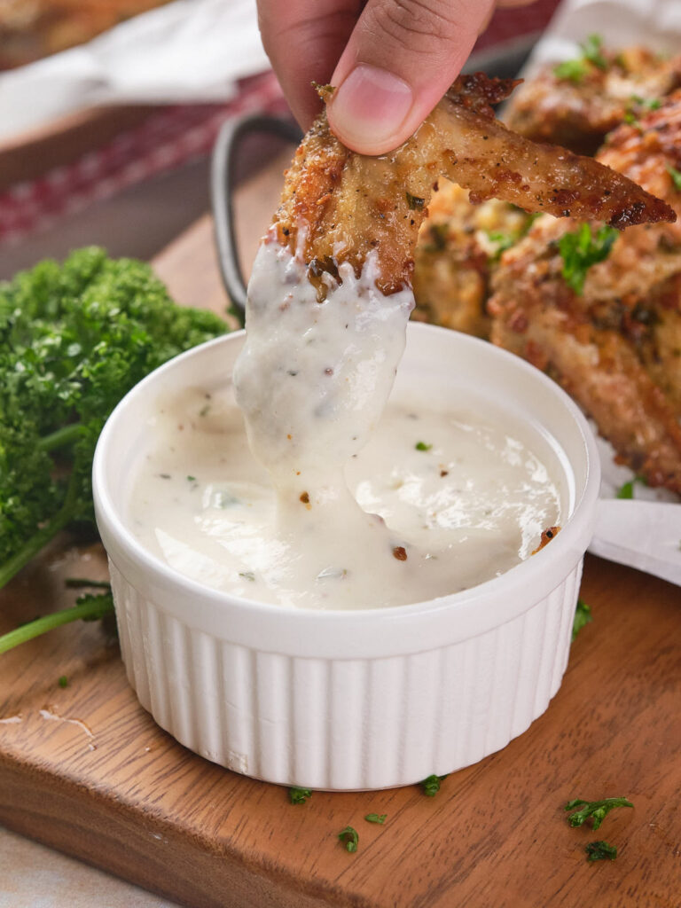 A hand dipping a seasoned chicken wing into a wingstop ranch dressing in a small white ramekin.