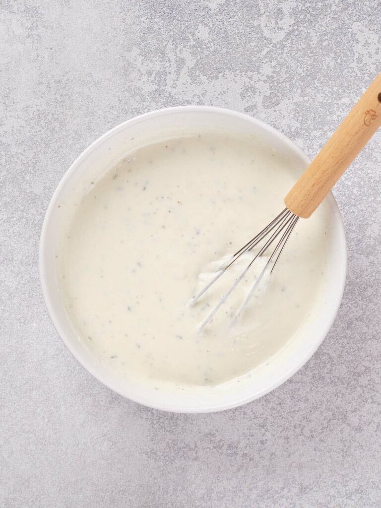 A bowl of wingstop ranch dressing with specks of herbs, being whisked with a wooden-handled whisk.