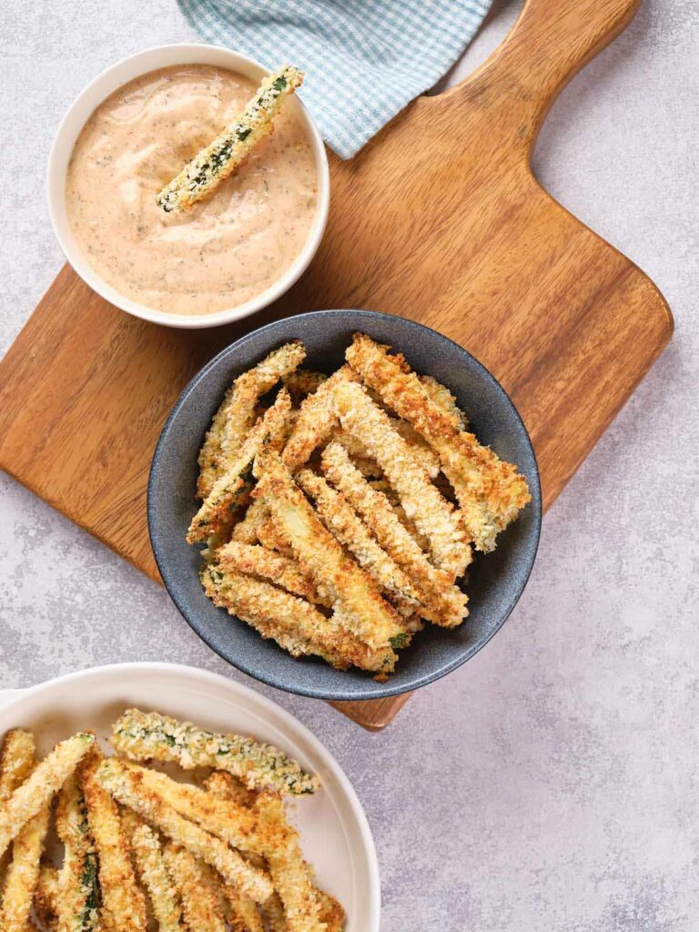 A bowl of breaded zucchini fries on a wooden cutting board with a small bowl of dipping sauce next to it.