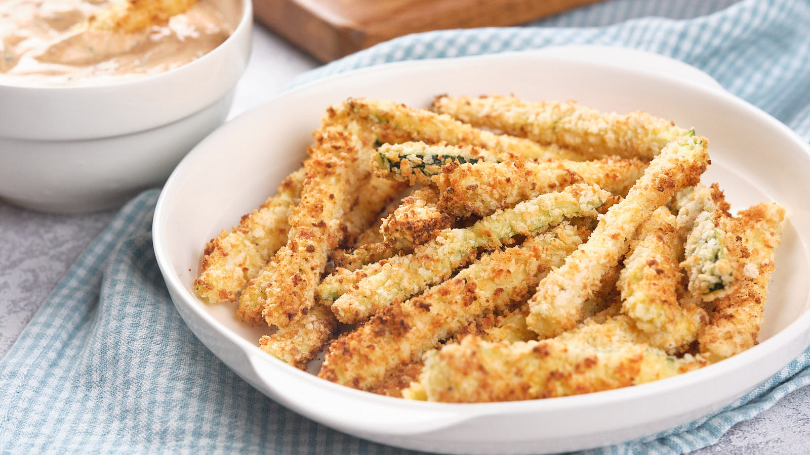 A white bowl filled with crispy, breaded air fryer zucchini fries is placed on a blue and white cloth.