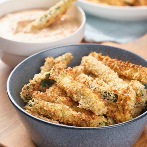 A bowl of crispy, breaded zucchini fries next to a bowl of creamy dipping sauce on a wooden surface.