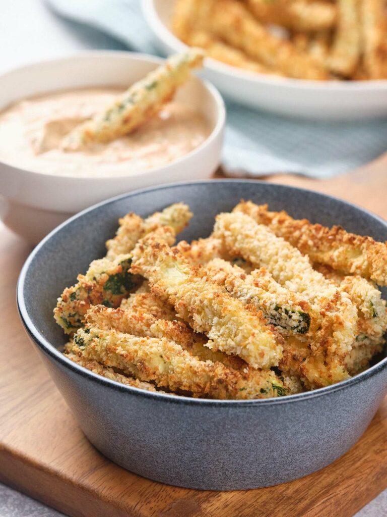 A bowl of crispy, breaded zucchini fries next to a bowl of creamy dipping sauce on a wooden surface.